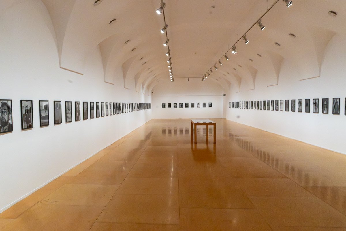 Could not look away_8 Long white gallery room with photographs lining both walls receding to a vanishing point, empty