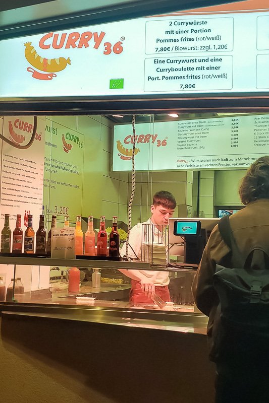 Europe H Counter worker at Curry 36 sausage stand in Berlin, menu boards and condiment bottles visible behind glass