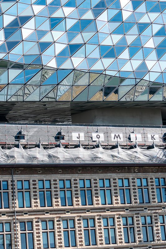 Europe H_16 Geometric glass diamond facade of the Port House in Antwerp rising above historic brick warehouse, partial letters J-o-M-i-K on scaffolding