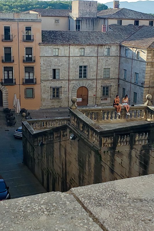Europe H_18 Two young women seated on a stone balustrade above Girona's old town plaza, cathedral steps and ochre buildings behind them