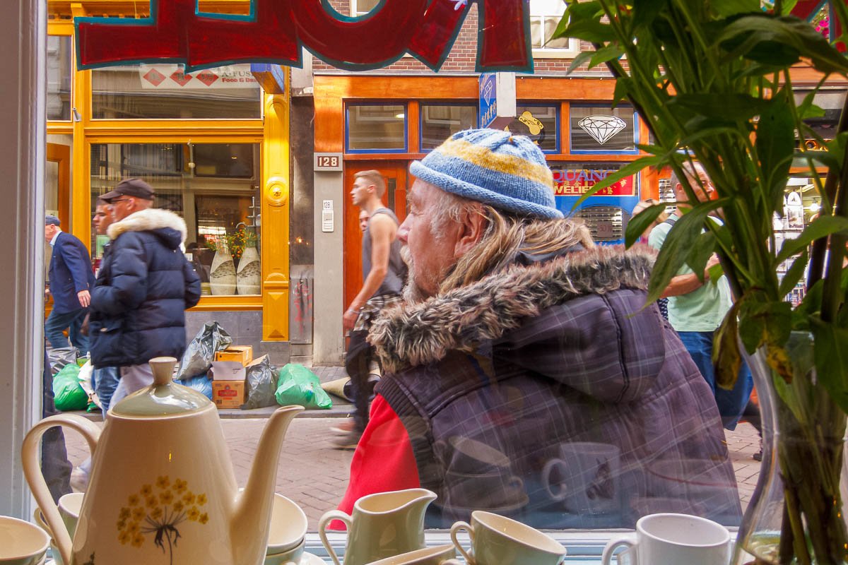 Europe H_4 Older man in knit cap seated at café window in Amsterdam, street life and teapots reflected in glass