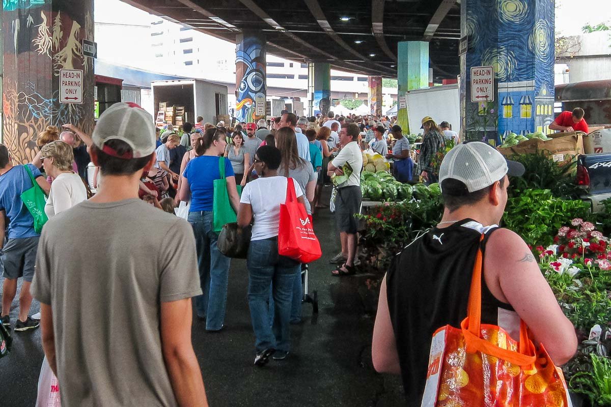 US_Housesit_18 Crowded Baltimore farmers market under a highway overpass, murals on concrete pillars, vendors and shoppers with produce and flowers