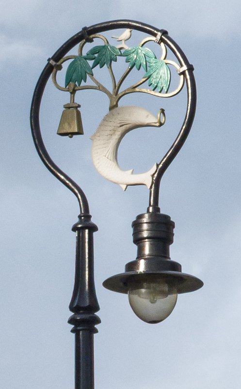 lights_1 Close-up of an ornate wrought-iron street lamp with a decorative moon and tree motif against pale sky