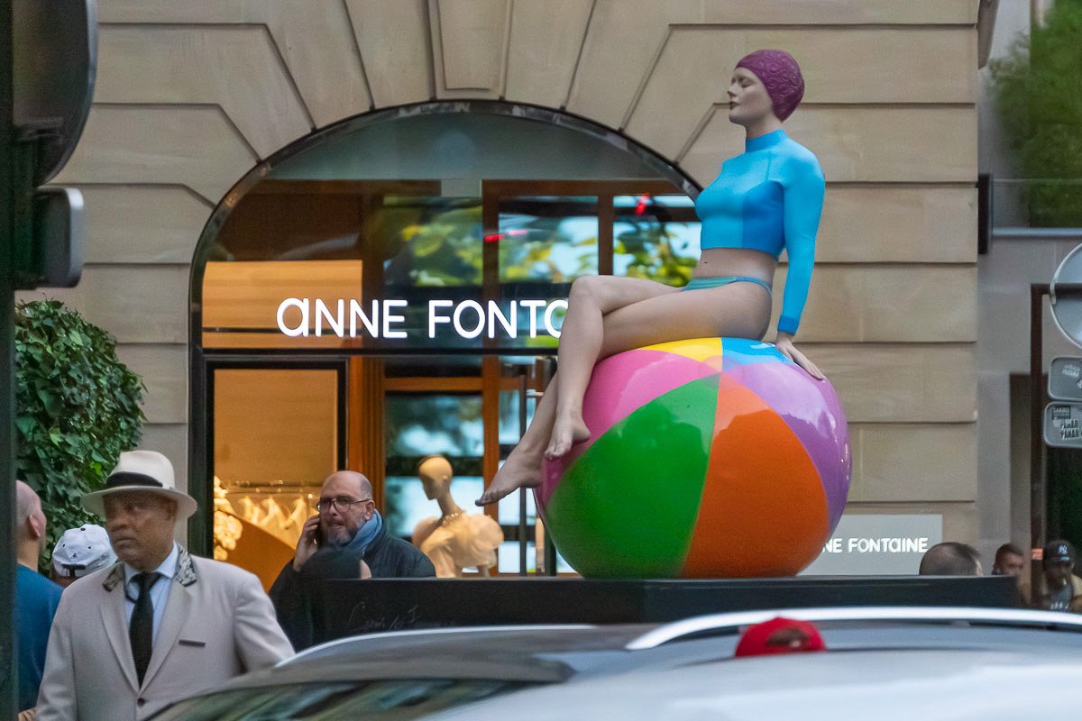 watchers_8 Street performer or sculpture of a clown balancing on a large ball outside a shop, absurdist